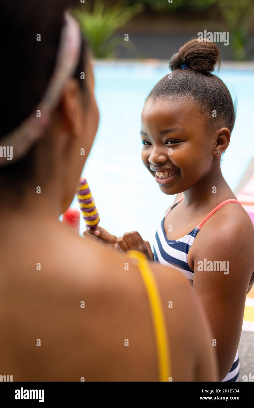 Happy african american mother and daughter sitting by pool in swimsuits holding ice creams Stock Photo