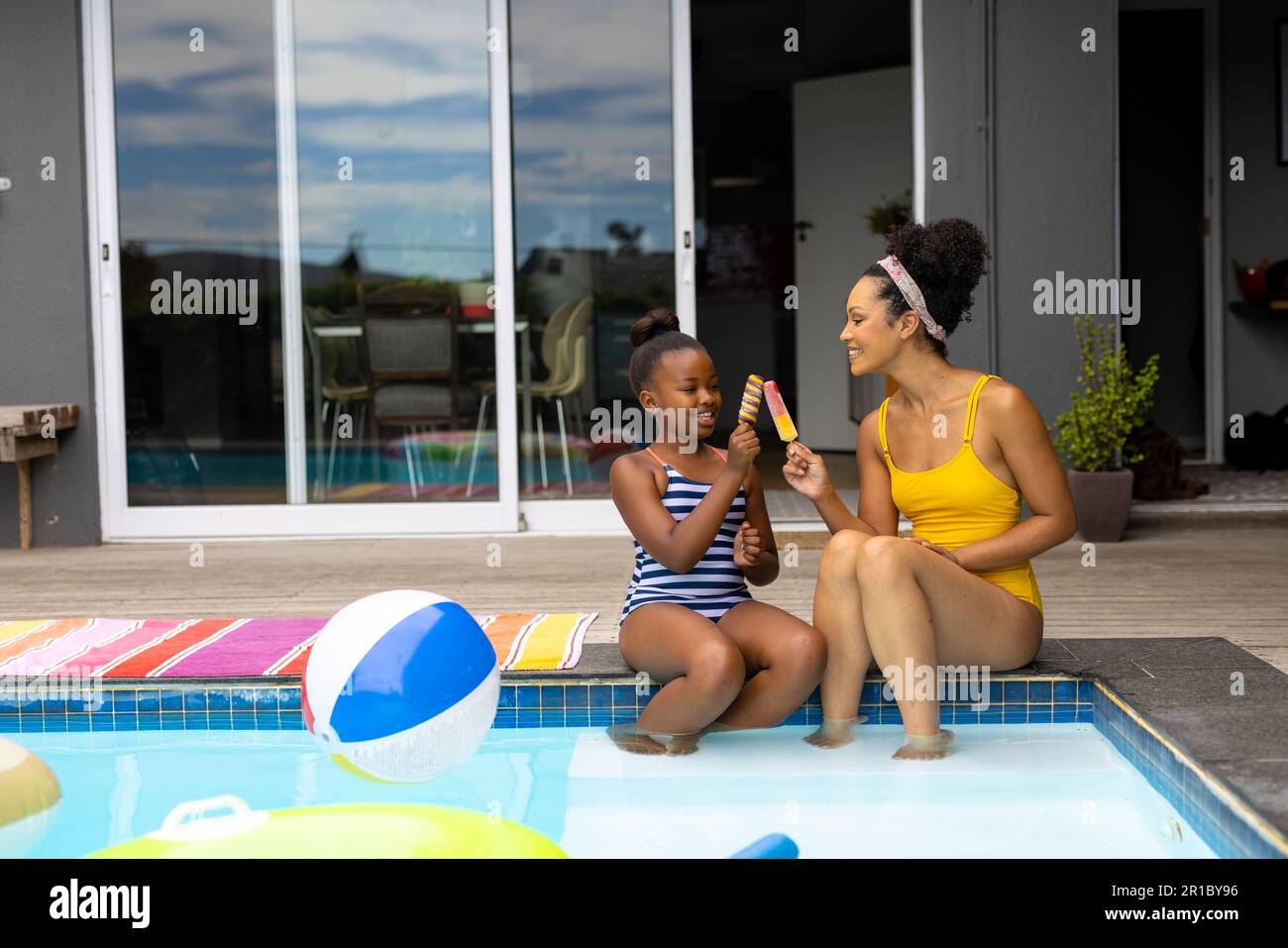 Happy african american mother and daughter by pool in swimsuits holding ice creams, copy space Stock Photo