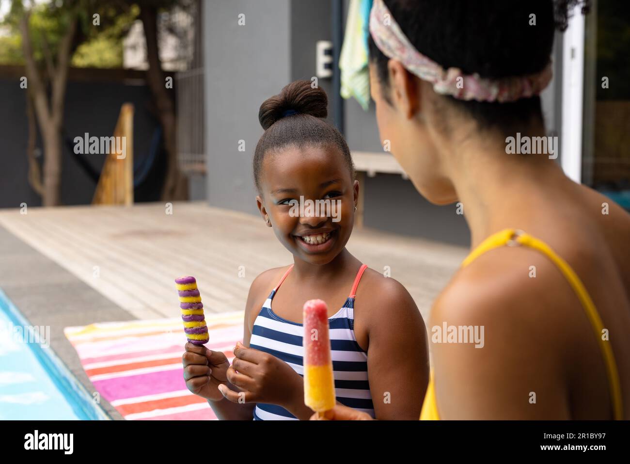 Happy african american mother and daughter sitting by pool in swimsuits with ice creams Stock Photo