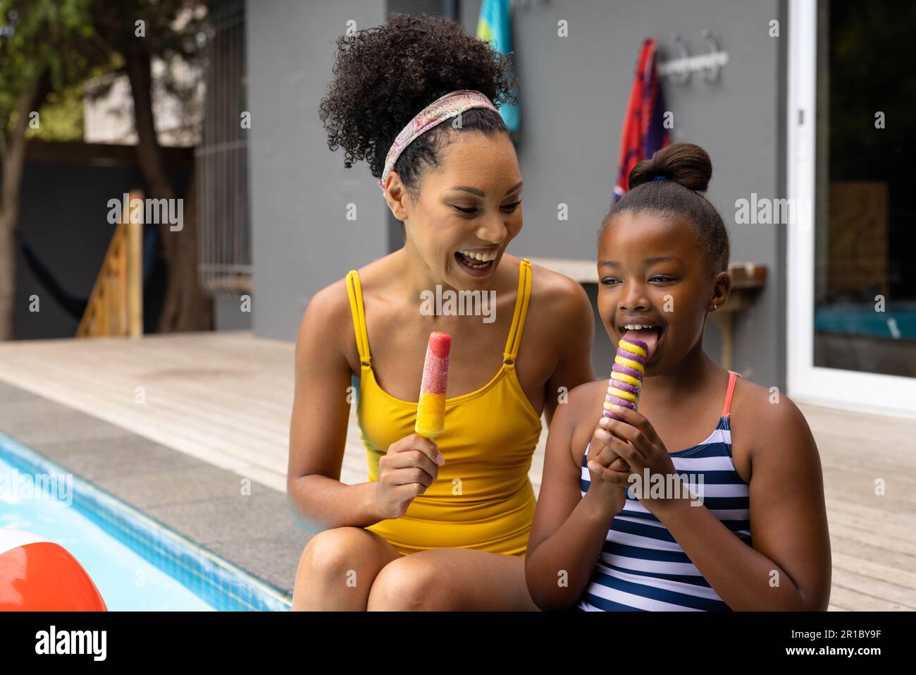 Happy african american mother and daughter sitting by pool in swimsuits eating ice creams Stock Photo