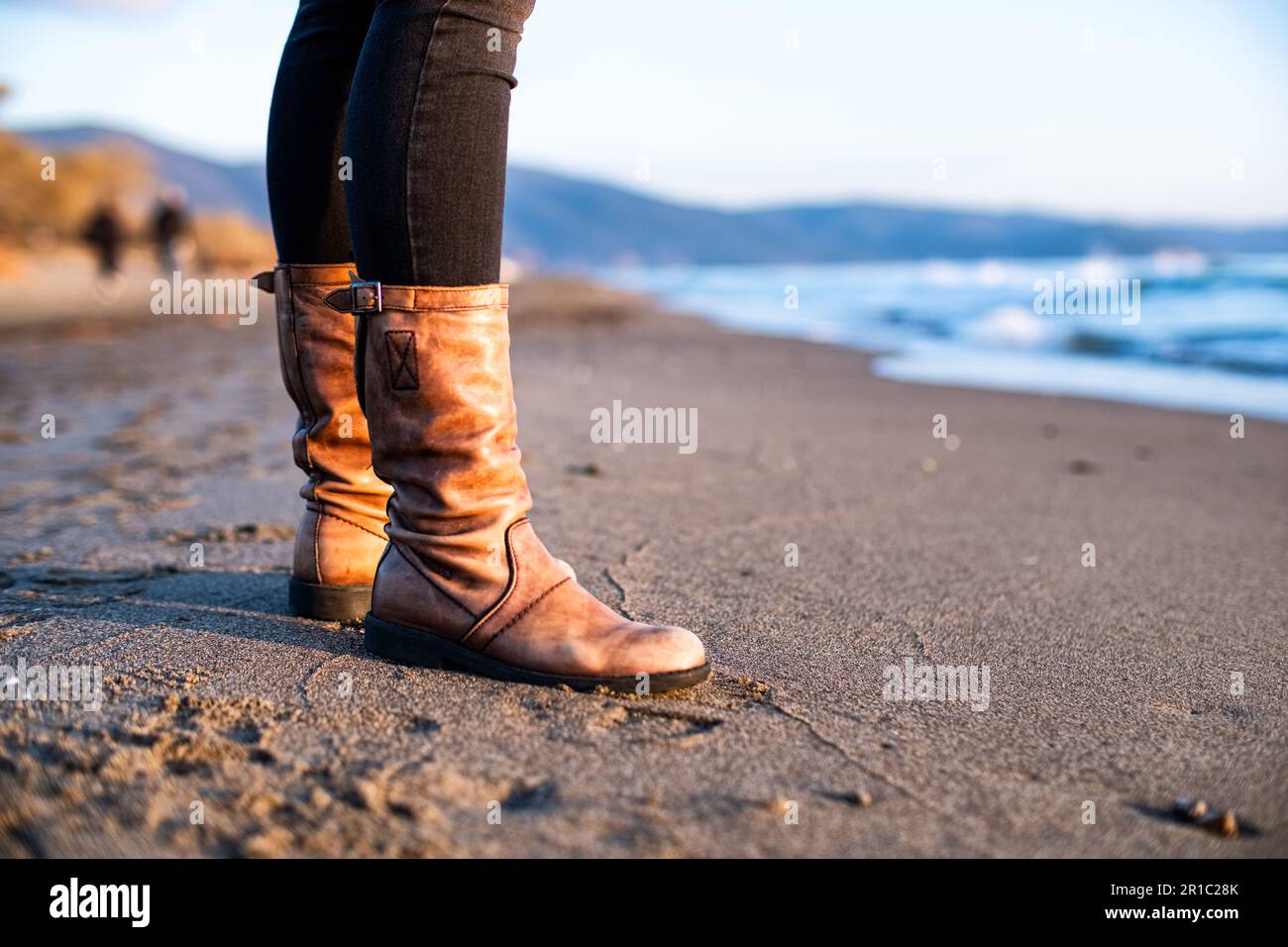 Sunset on the beach. A woman wearing orange leather boots, standing up on the beach sand. Mediterranean Sea in the foreground. Marina Di Grosseto. Stock Photo