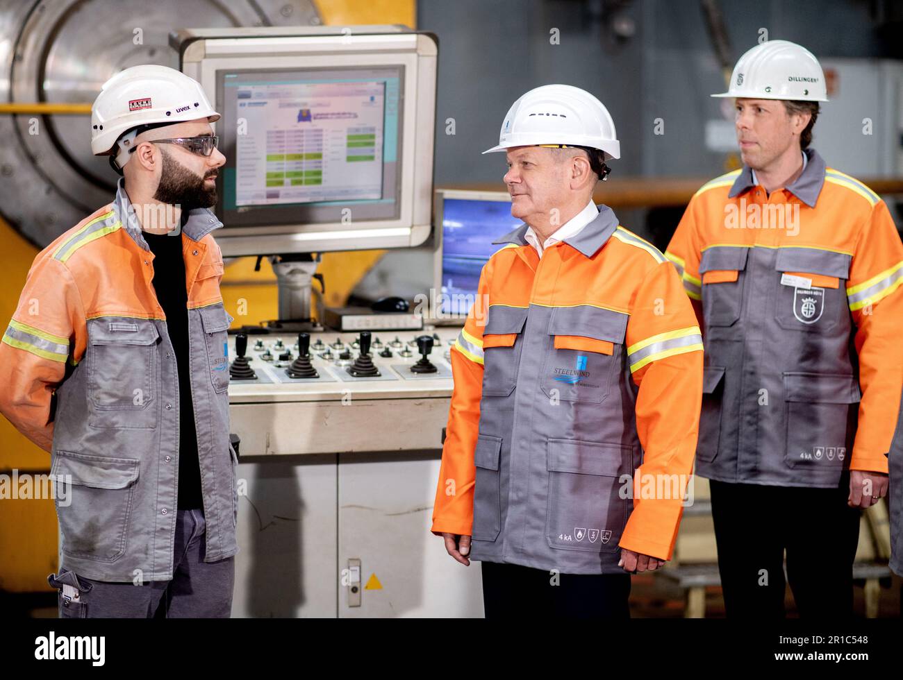 Nordenham, Germany. 12th May, 2023. Adem Dener (l-r), a production employee at Steelwind Nordenham GmbH, explains to German Chancellor Olaf Scholz (SPD) and Jonathan Weber, member of the Executive Board of Dillinger Hüttenwerke AG, the function of a bending machine for the production of steel foundations used for wind turbines. Scholz visited the production of steel foundations, so-called monopiles, for wind turbines in offshore wind farms at Steelwind Nordenham GmbH. Credit: Hauke-Christian Dittrich/dpa/Alamy Live News Stock Photo