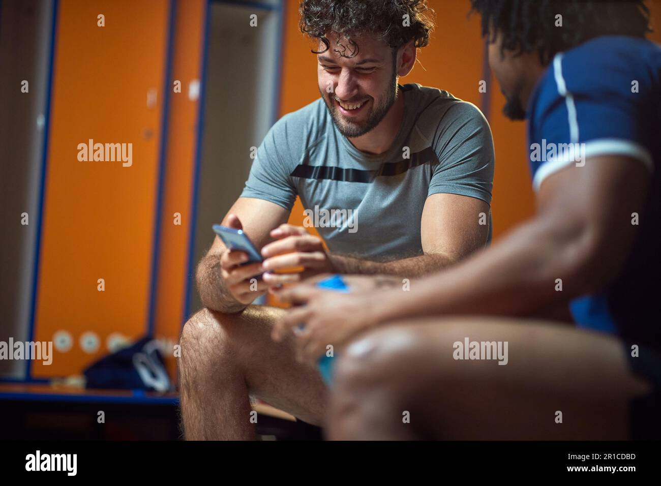 Young man using mobile phone with friend sitting on bench in the gym locker room, having fun before workout. Sports, health, togetherness concept. Stock Photo