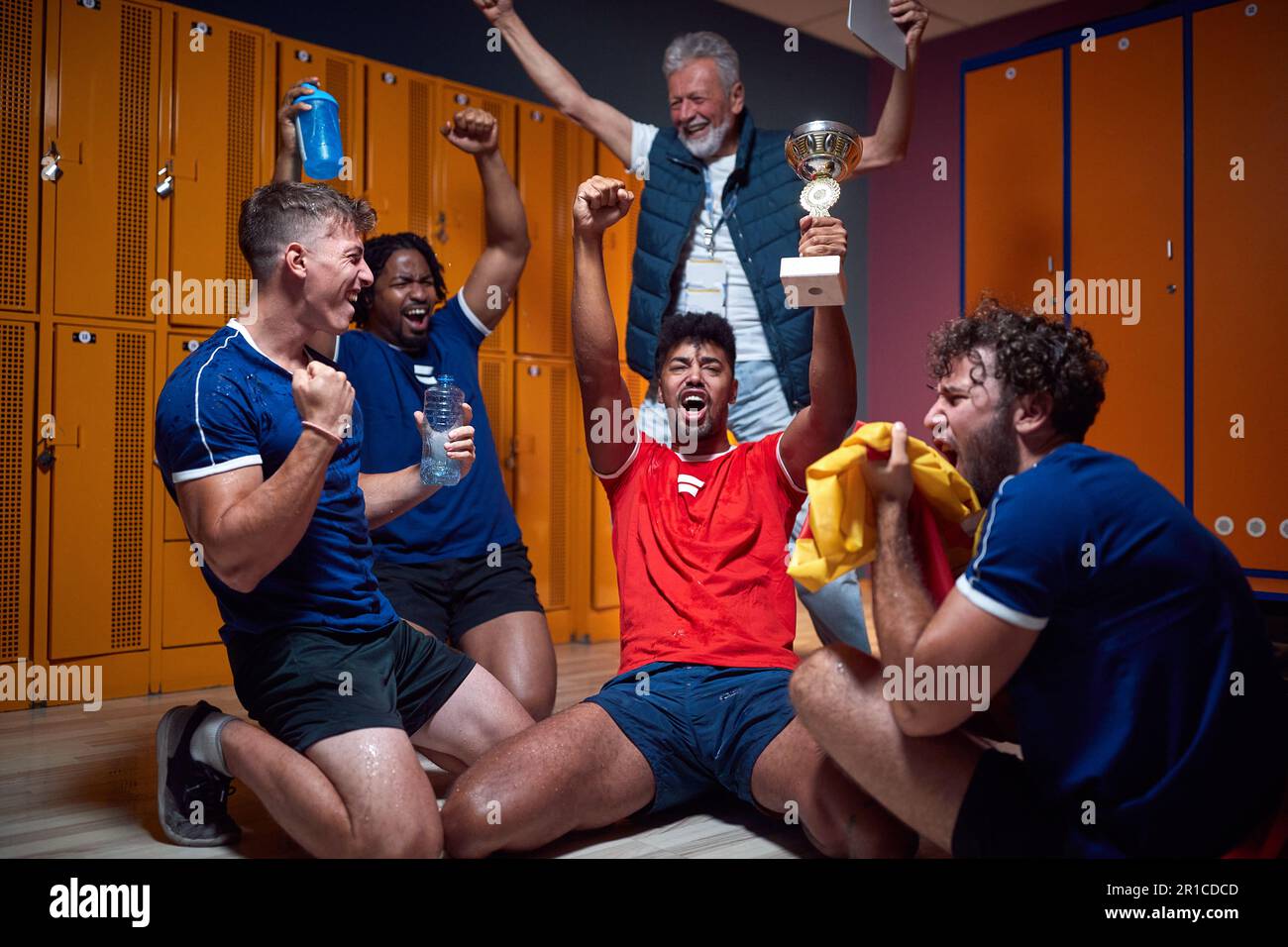 Group of young football players celebrating success in the locker room with their senior coach in the background. Sports, active lifestyle concept. Stock Photo
