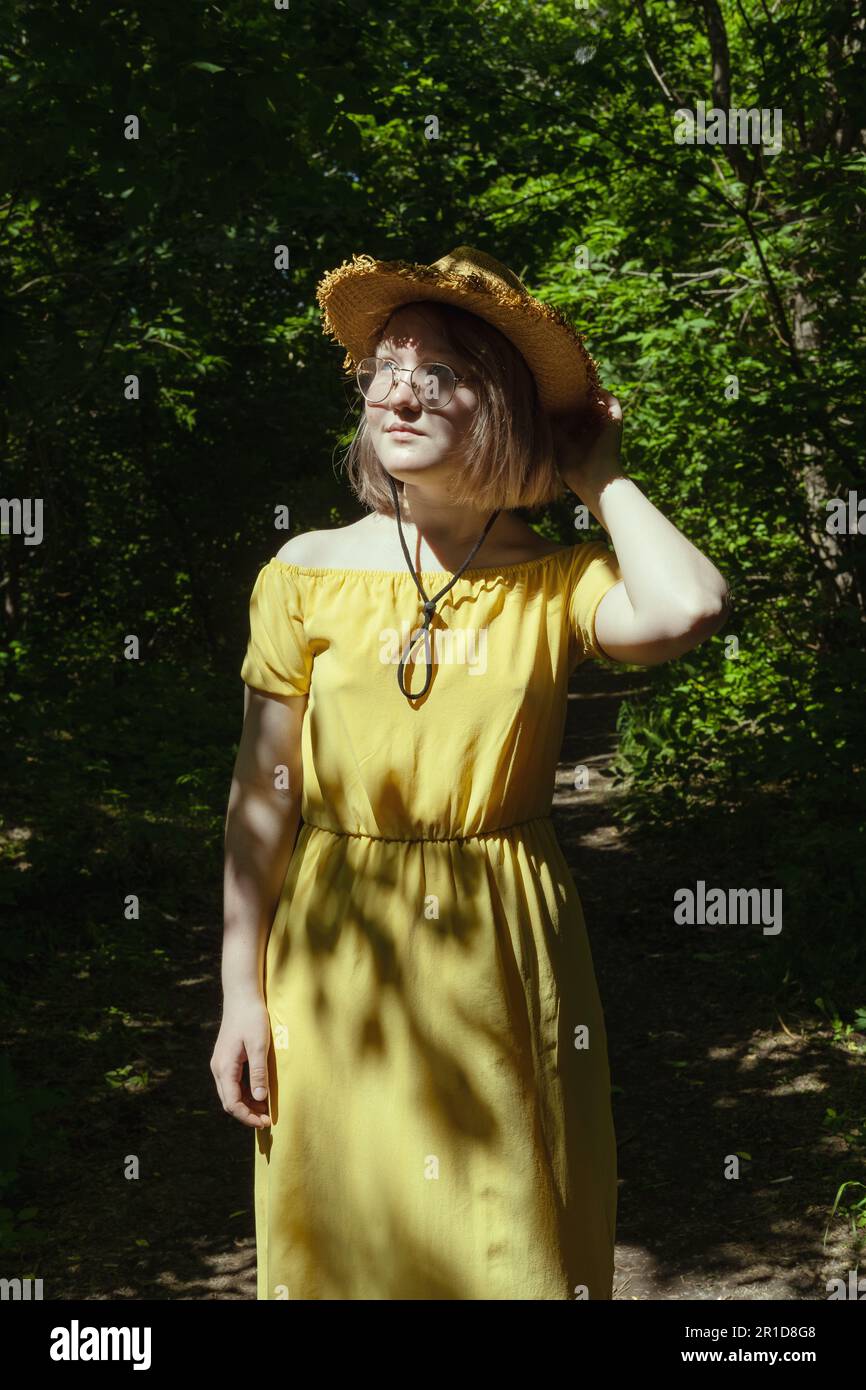 Beautiful Asian girl with glasses, straw hat and yellow dress in the forest in summer, beautiful shadows, sunlight, portrait. Dreamy young teen girl. Stock Photo