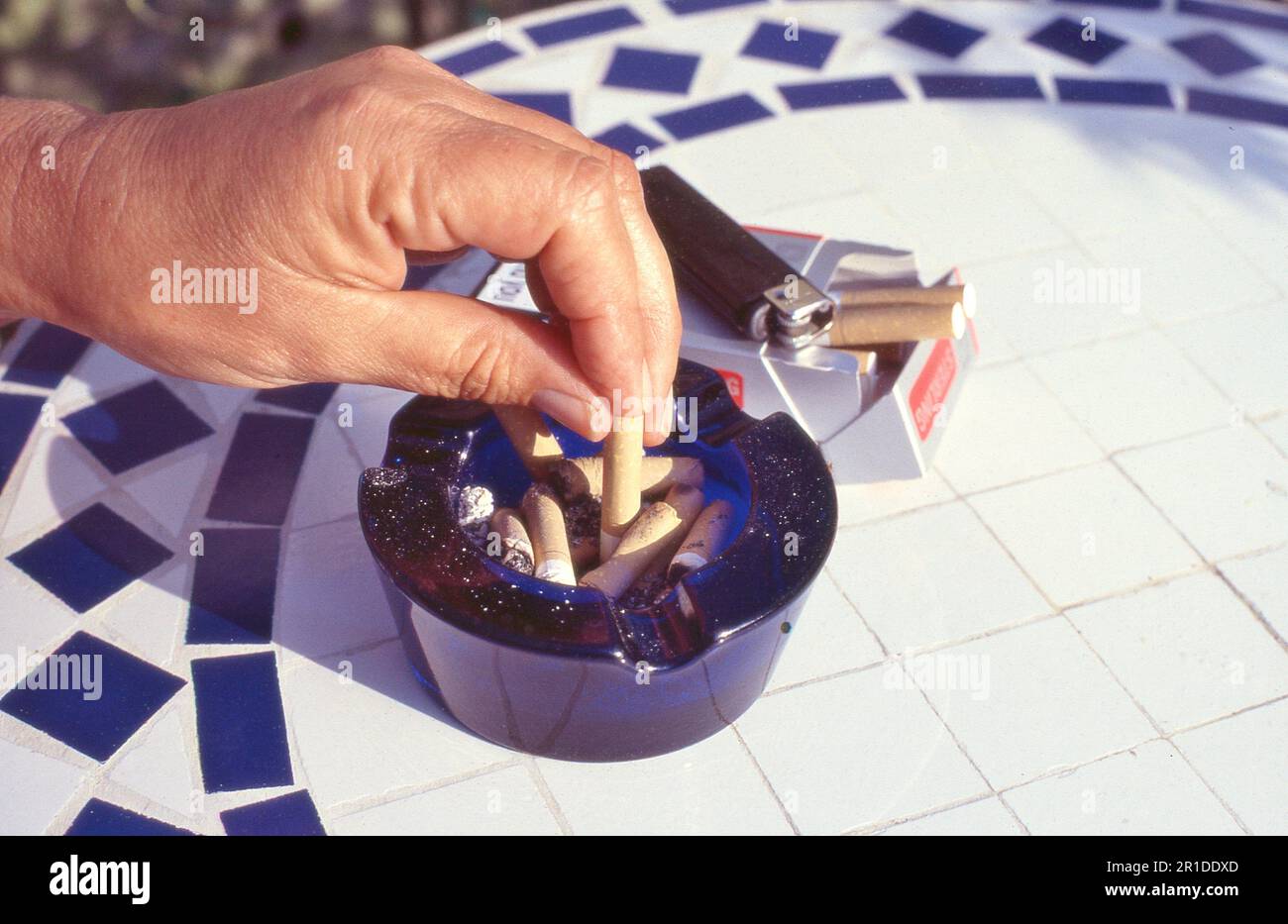A woman stubs out a cigarette into a full ashtray with a packet of cigarettes and lighter nearby. Stock Photo