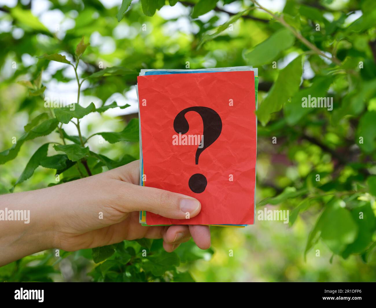 A woman holding a stack of colorful paper notes with question marks on them in her hand against a background of foliage. Close up. Stock Photo