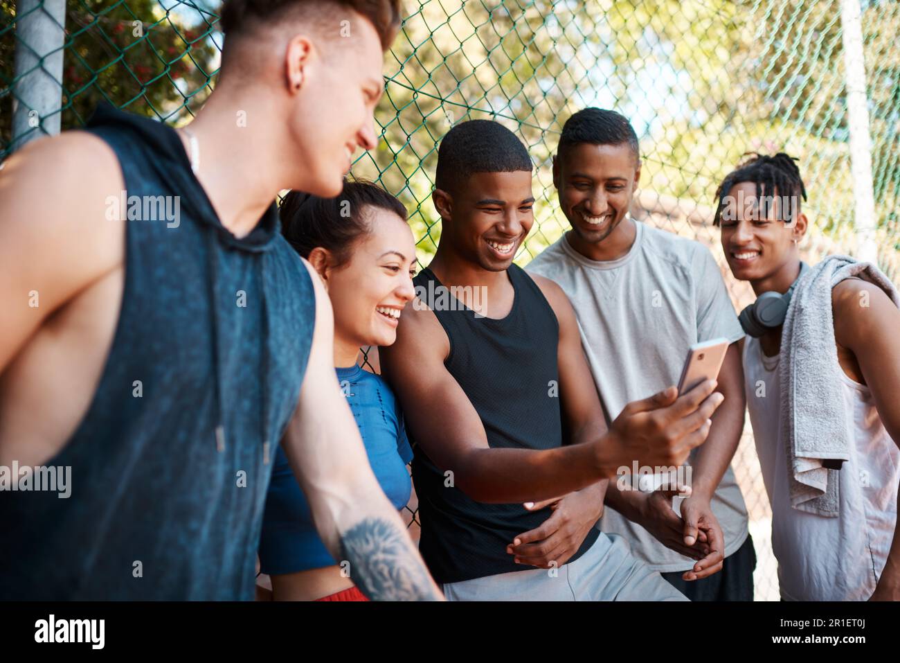 Told you this would be hilarious. a group of sporty young people looking at something on a cellphone while standing along a fence outdoors. Stock Photo