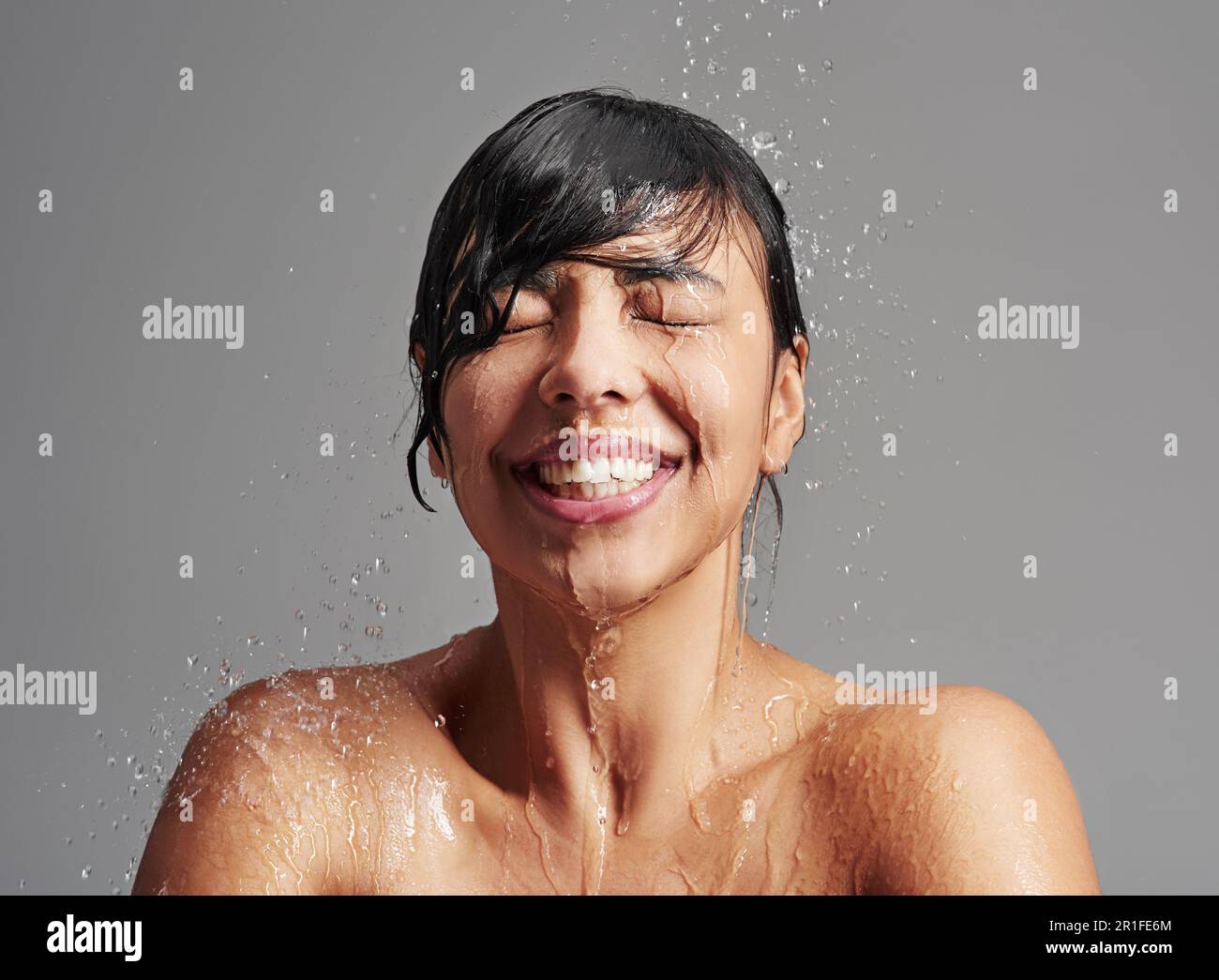 Shower, water splash and face of woman in studio on gray background for wellness, cleaning and