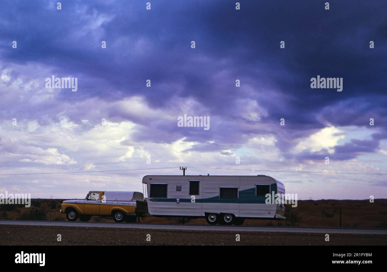 Pick up truck pulling a camper trailer under a stormy sky ca. 1964