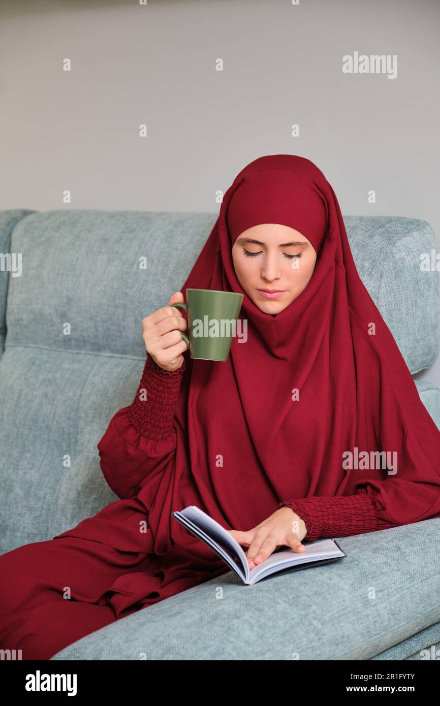 Muslim young woman in hijab reading a book sitting on sofa at home ...