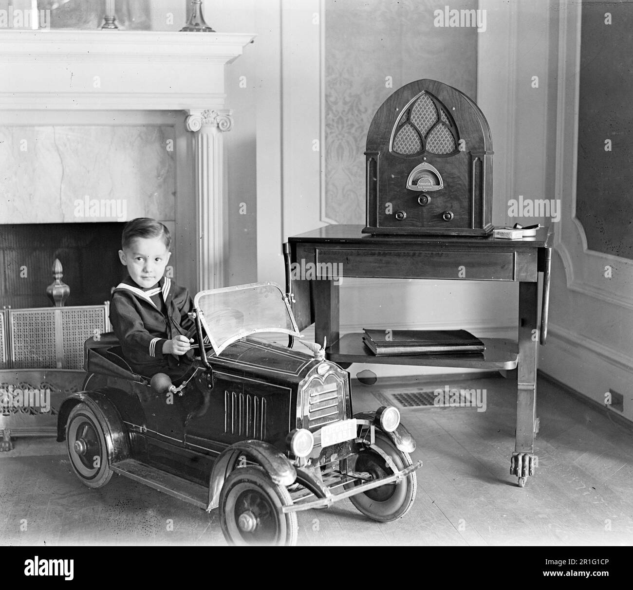 Archival Photo: Young boy playing in a toy automobile in a living room, with radio ca. 1910s or 1920s Stock Photo