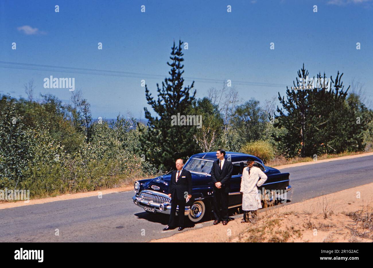 Two well dressed executives and their driver outside of their automobile, parked alongside a road in South Africa, possibly Capetown area ca. 1961 Stock Photo