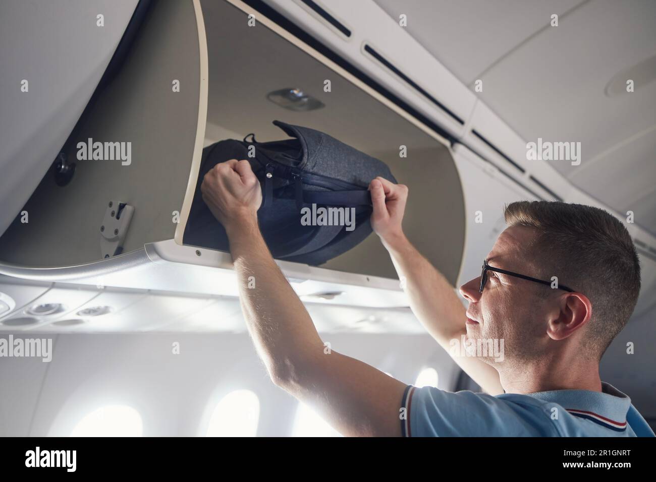 Man travel by airplane. Passenger putting hand baggage in lockers above seats of plane Stock