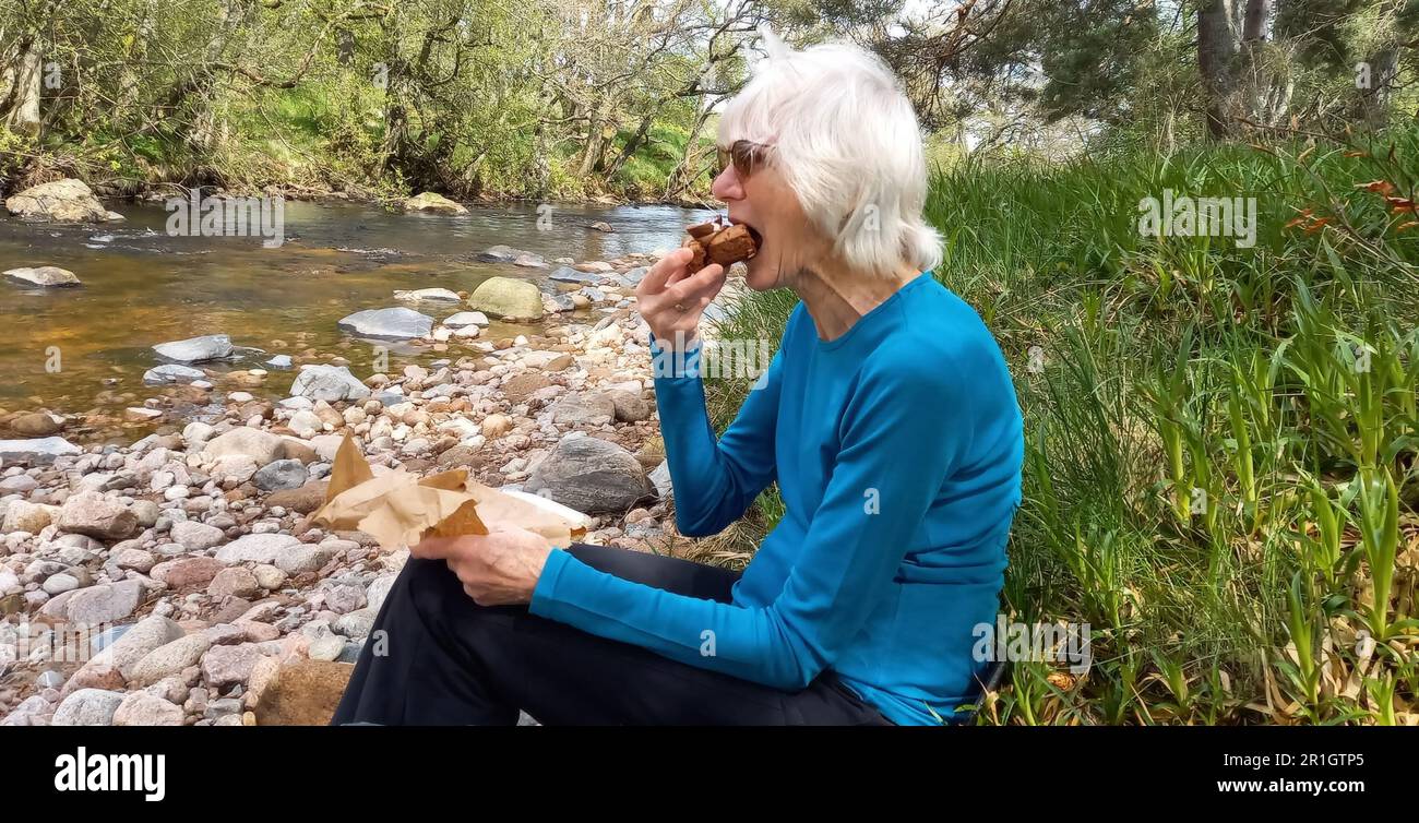 Senior lady resting and eating chocolate brownie following a long walk Stock Photo
