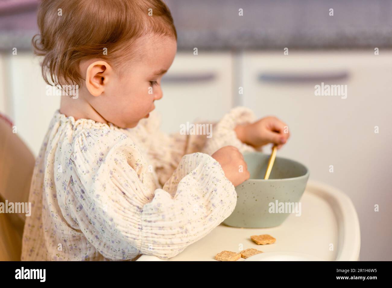 Little baby girl sitting in a highchair eating by herself with a spoon Stock Photo