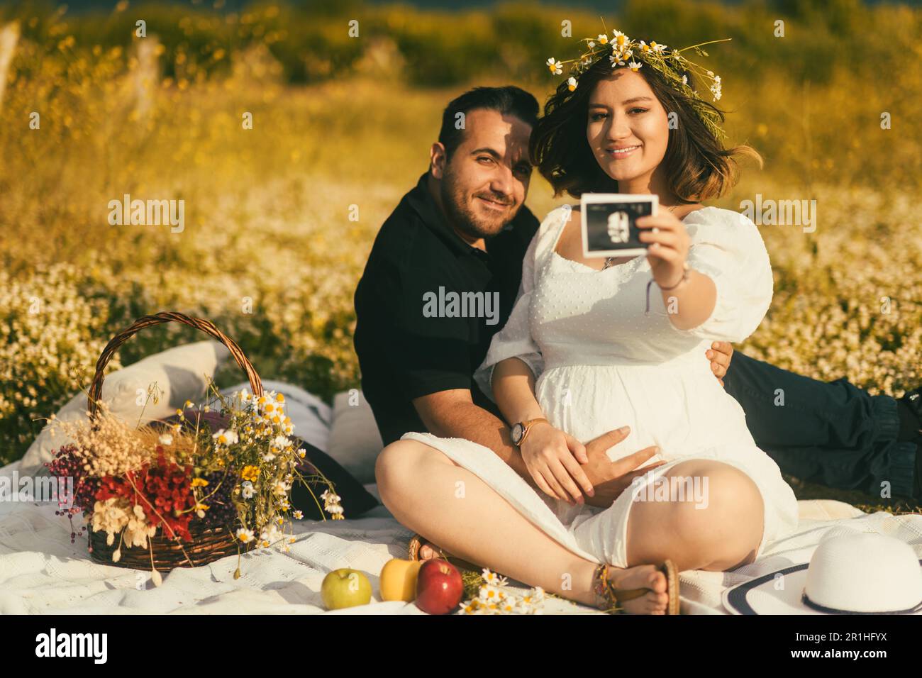 Sitting on a white picnic blanket, a pregnant woman with a daisy crown and her husband are sharing a joyful moment, he touches her belly while she pro Stock Photo