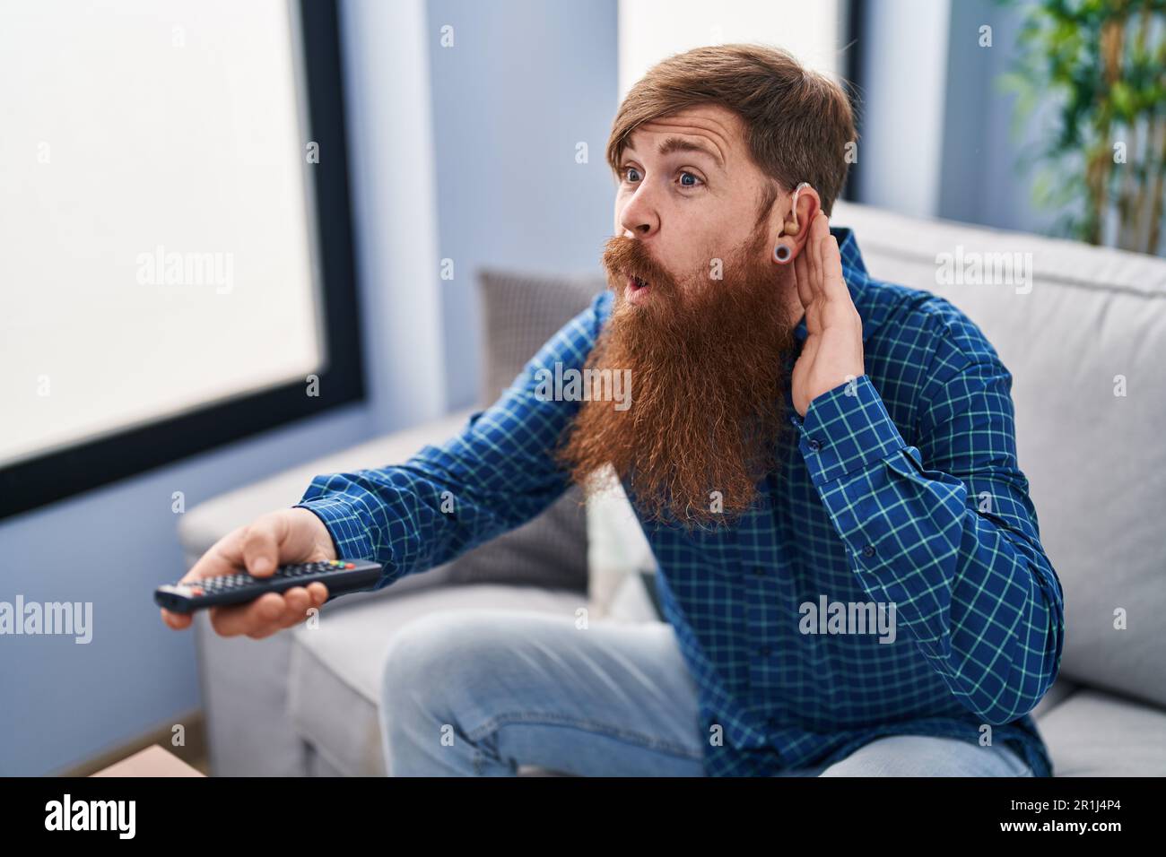 Young redhead man using deafness aid machine watching tv at home Stock Photo