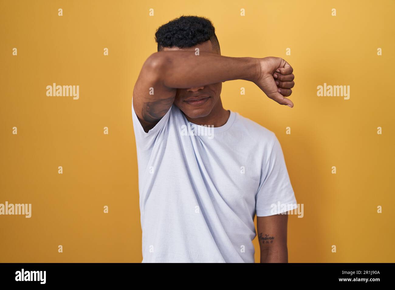 Young hispanic man standing over yellow background covering eyes with ...