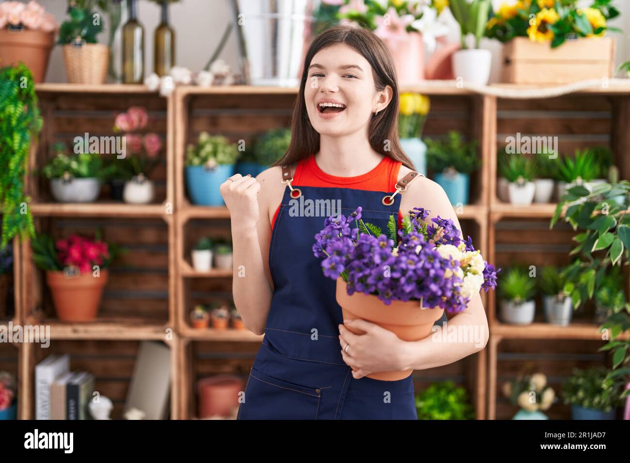Young caucasian woman working at florist shop holding pot with flowers screaming proud, celebrating victory and success very excited with raised arms Stock Photo