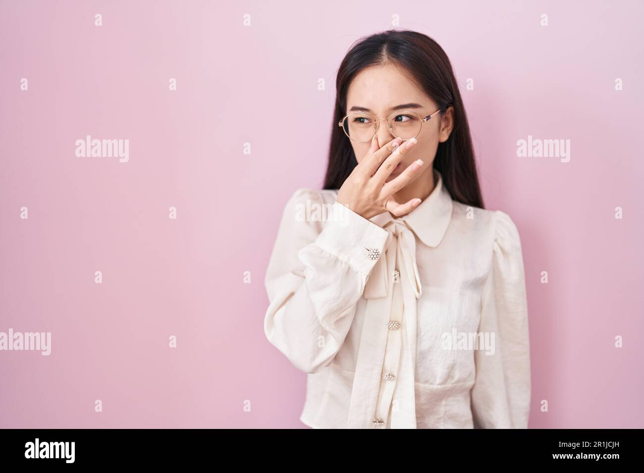 Young chinese woman standing over pink background smelling something stinky and disgusting, intolerable smell, holding breath with fingers on nose. ba Stock Photo