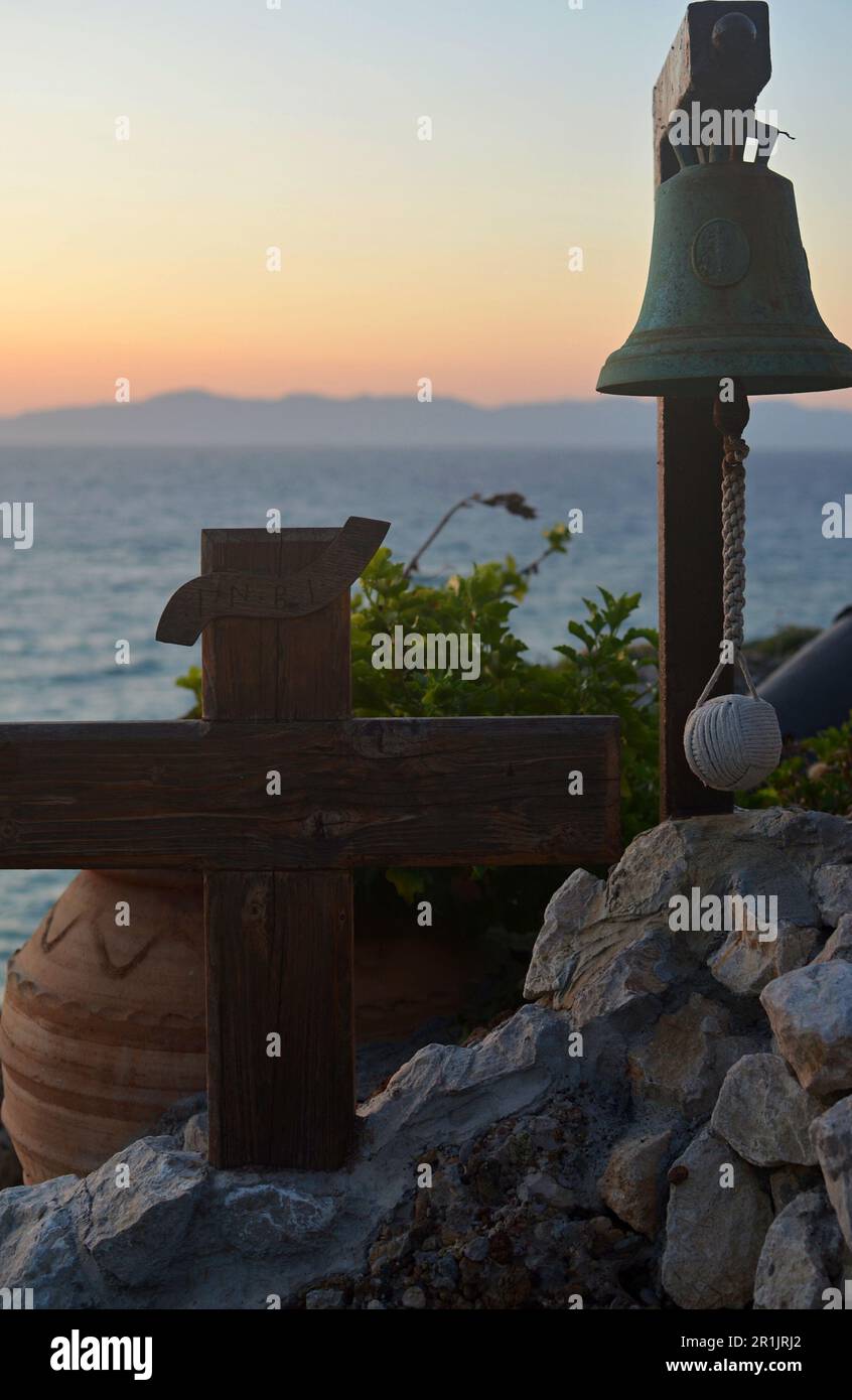Wooden Orthodox cross and a small bell on a wooden stand on a rocky hill overlooking the sea and sunset Stock Photo