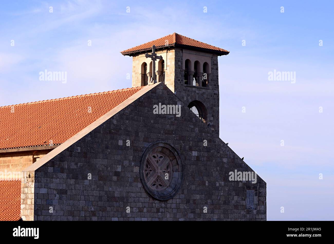 Catholic church in the Gothic style with a bell tower, a cross and a red roof. Trees by the road