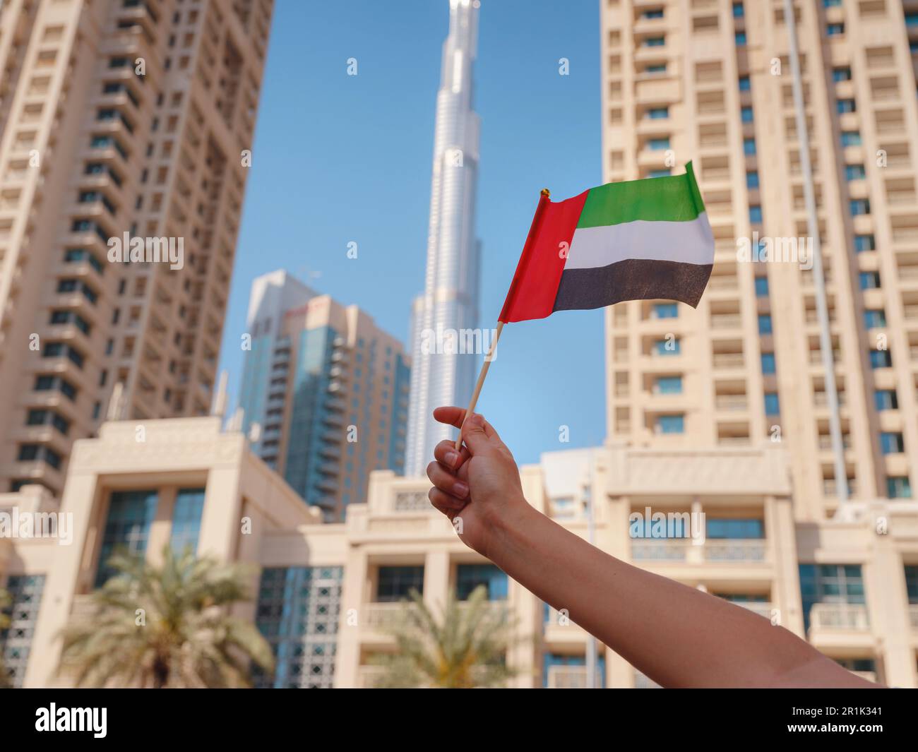 Close up of a female hand holding tiny flag of UAE against Dubai skyline. Photo of UAE national day celebration spirit. Stock Photo