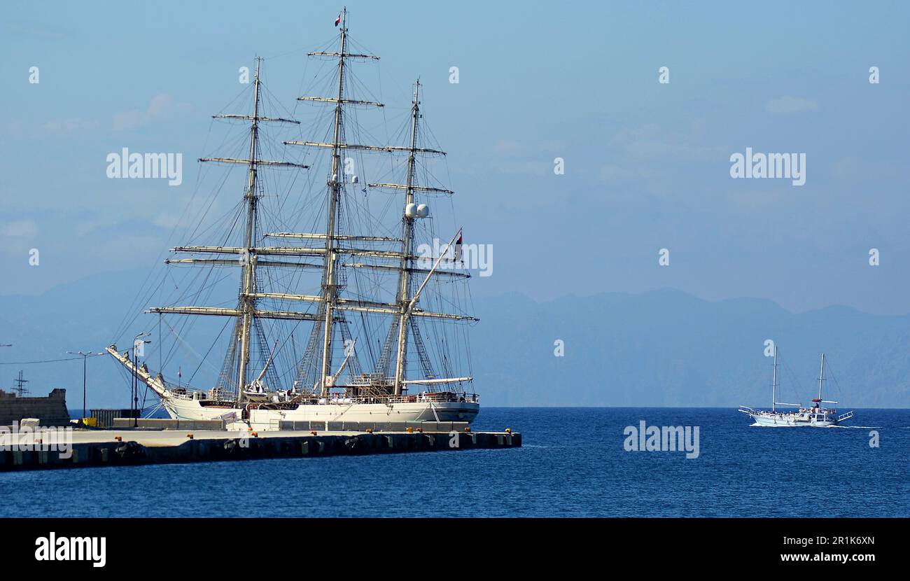 A white three-masted sailboat with many sails is moored in the port of the island of Rhodes. Frigate with assembled sails near the embankment. Stock Photo