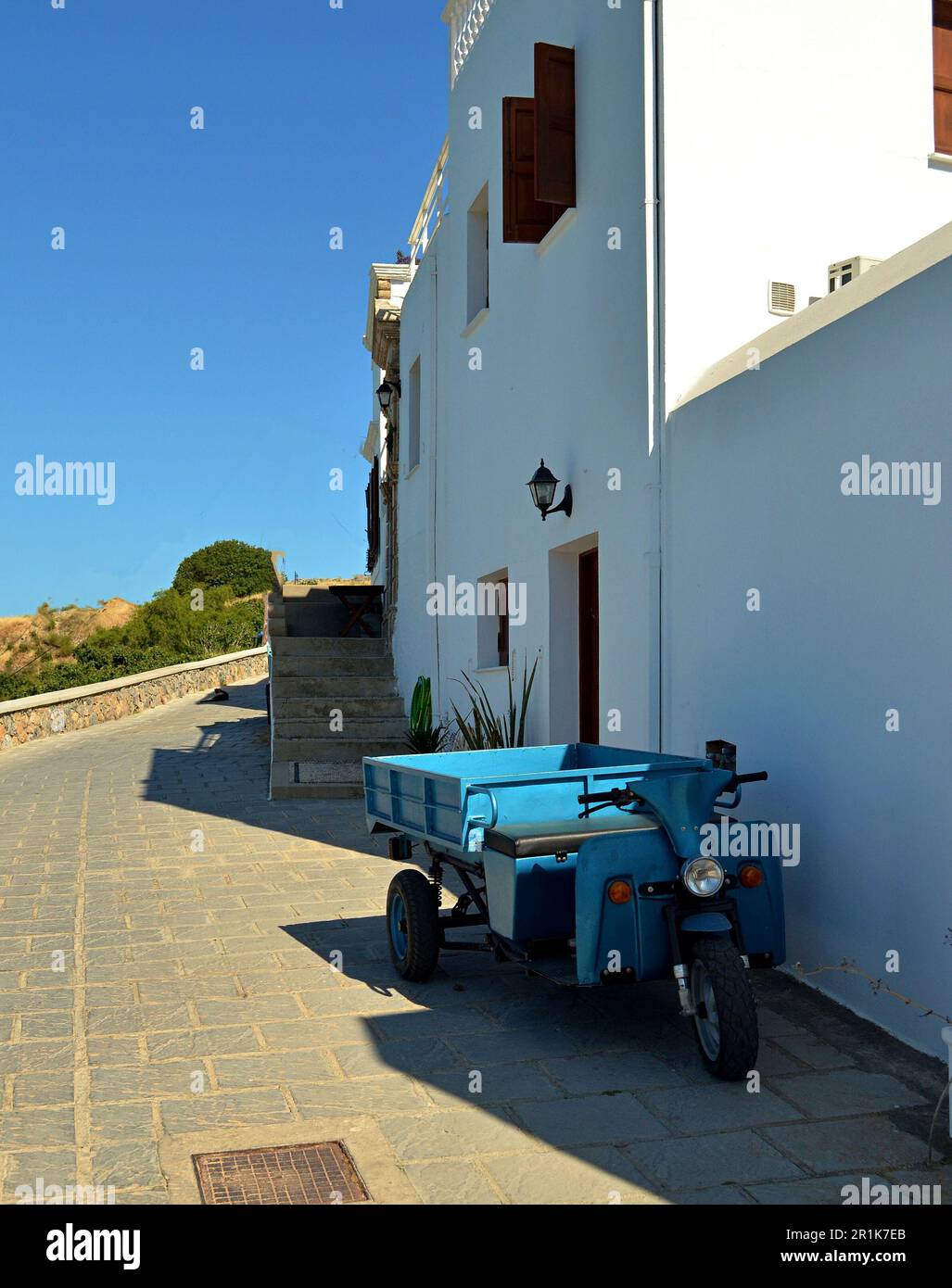A blue old motorcycle with a sidecar is parked on a village street in Lindos. Near the white walls of a house with wooden shutters and a door. Stock Photo