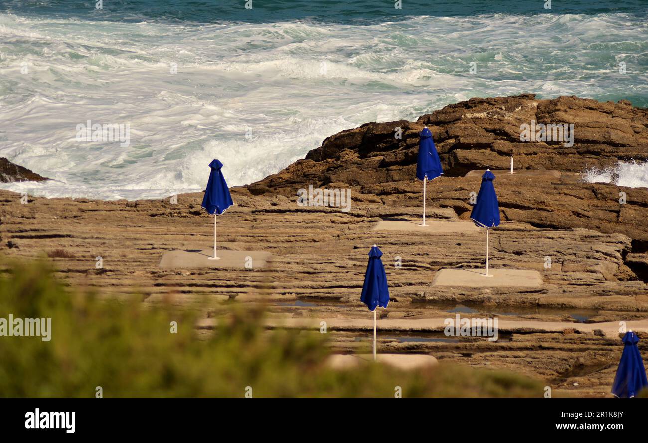 Coast of rocky beach in winter. Strong white sea waves, storm, umbrellas closed. For each umbrella there is a concrete platform. Stock Photo