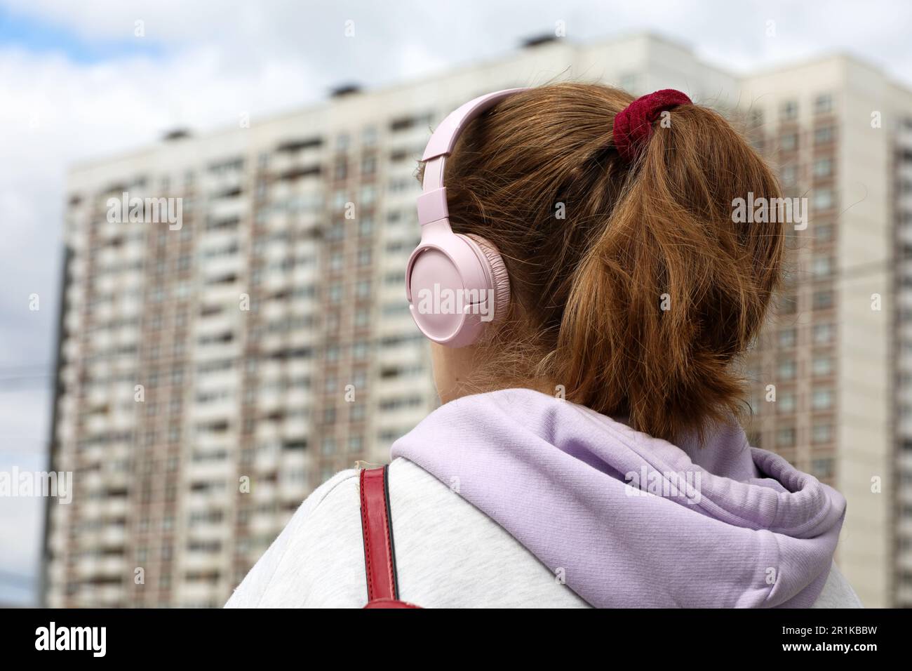 Girl with pink headphones standing on residential buildings background. Headset, listening to music and city life concept Stock Photo