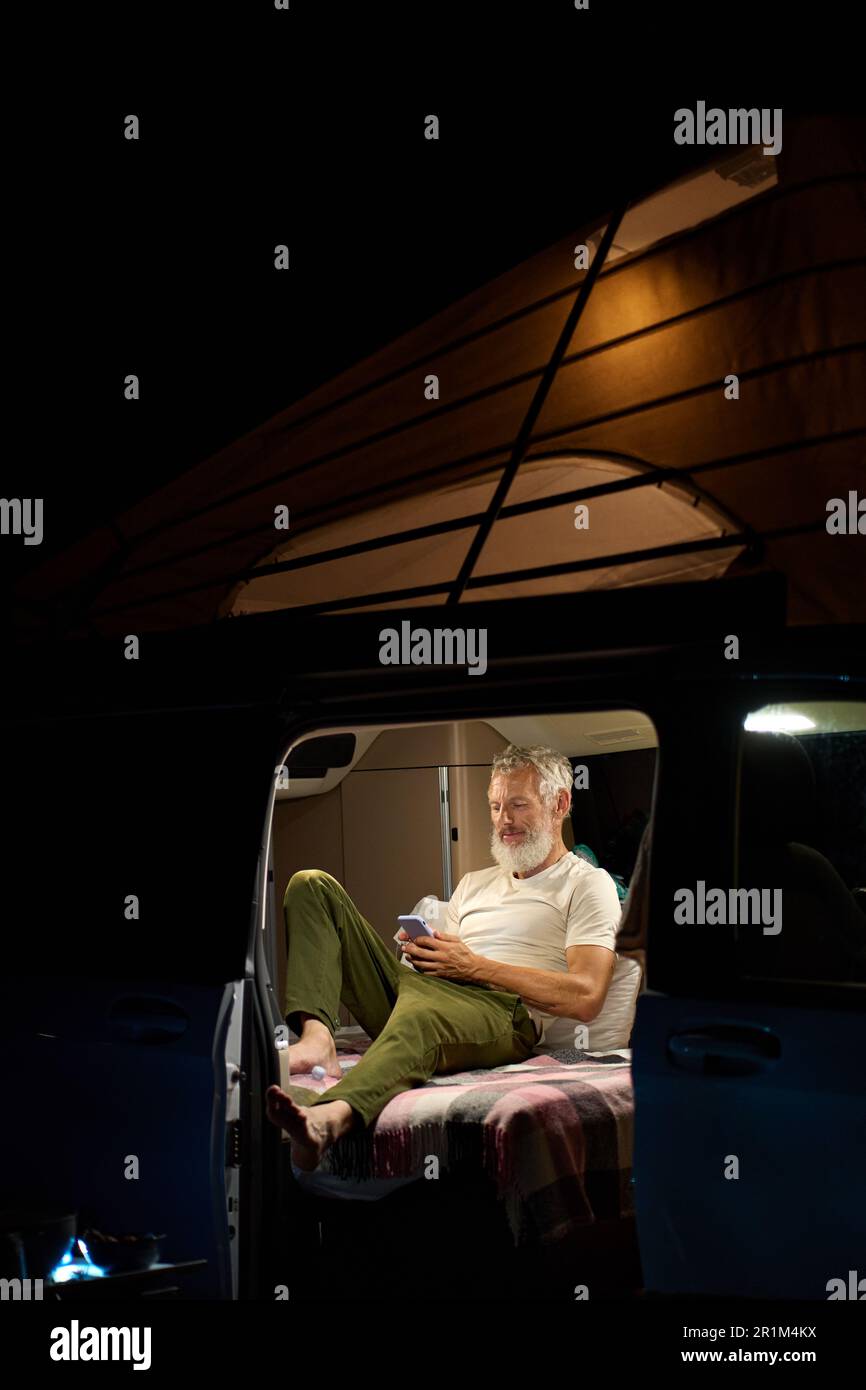 Older man traveler sitting in camper van using mobile in camping at