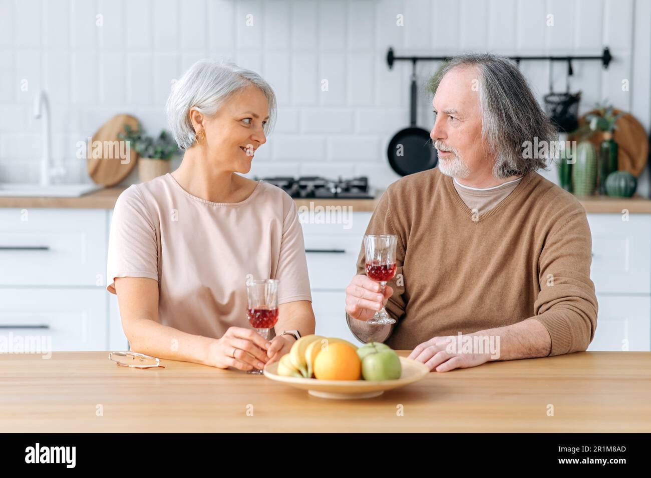 Middle aged caucasian spouses, sit at home in the kitchen, hold glasses of red wine in their hands, celebrate an important date, anniversary, look at glasses, smile joyfully. Happy pensioners Stock Photo