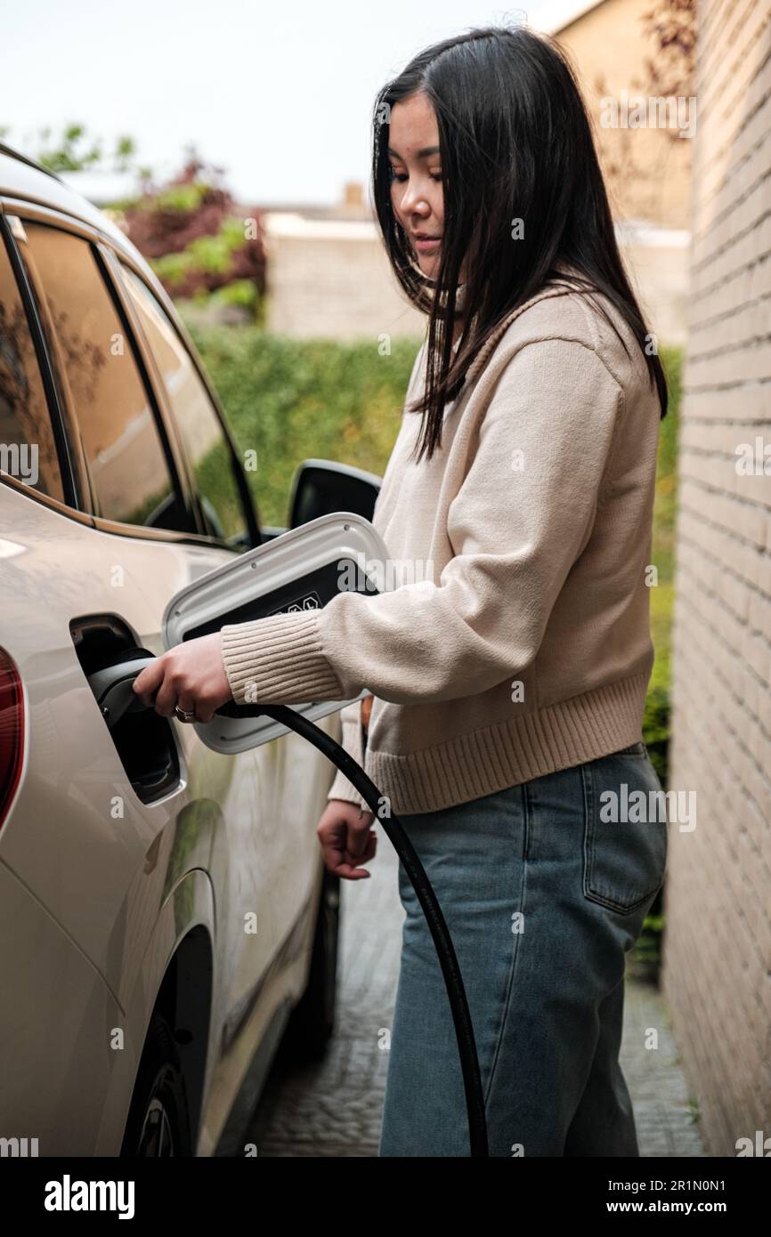 Young Central Asian woman plugging in a charging cable to charge her electric car. Sustainable and economic transportation concept. Stock Photo