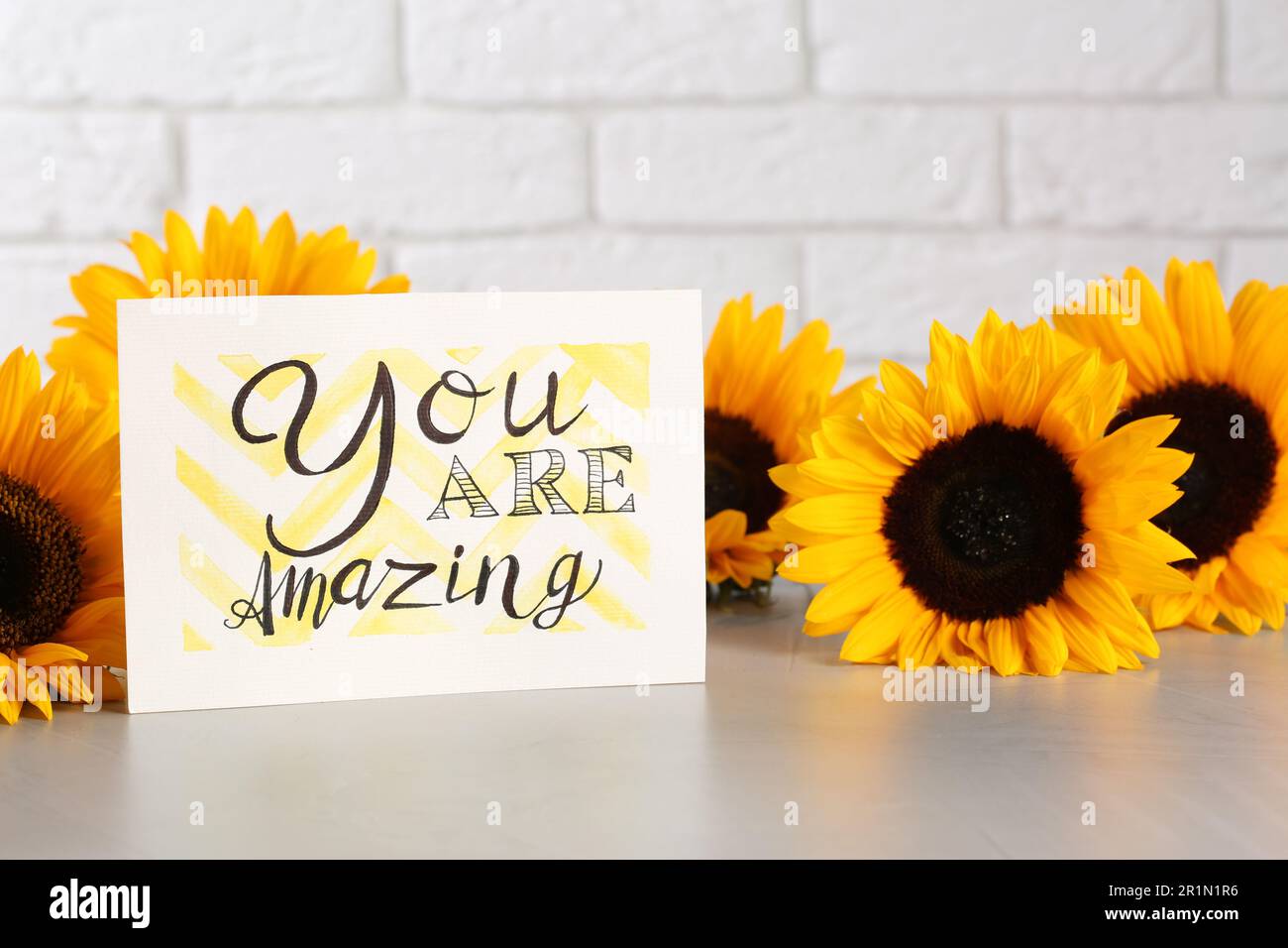 Card with life-affirming phrase You Are Amazing and sunflowers on light table against white brick wall Stock Photo