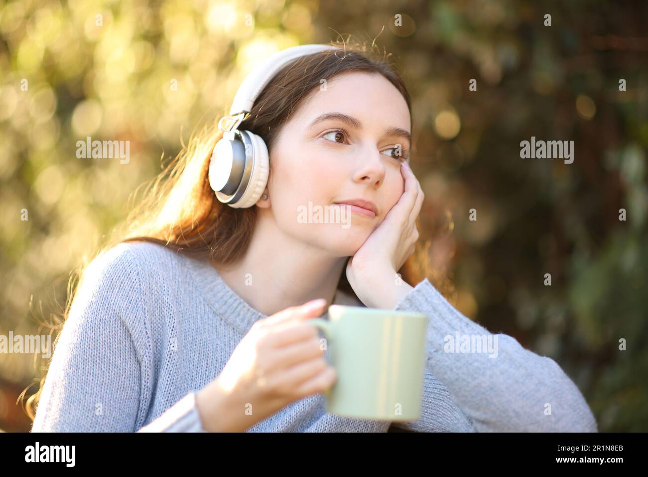 Pensive woman holding coffee and listening to music looking away in a park Stock Photo