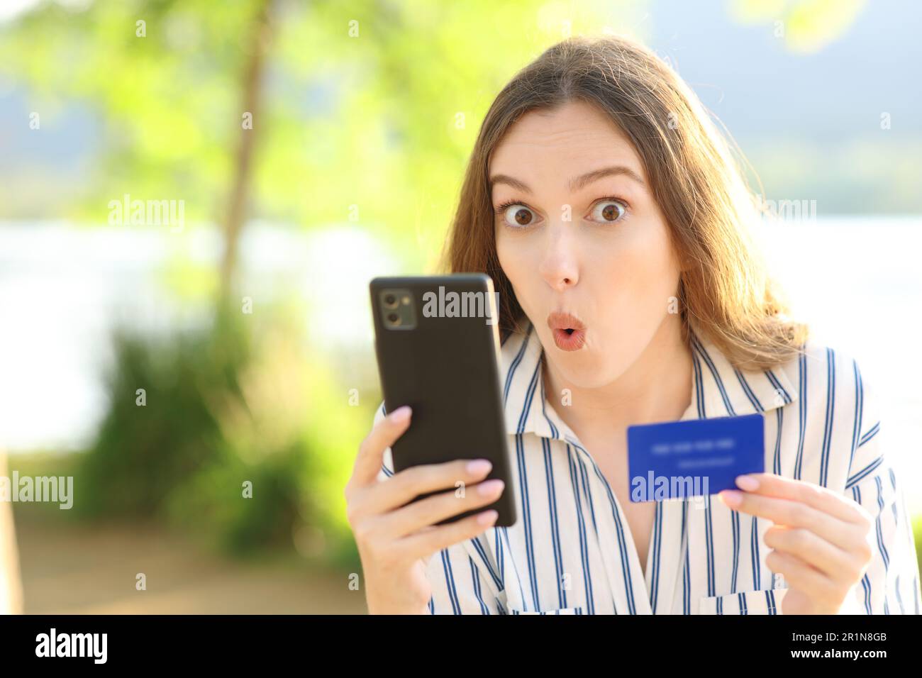 Surprised online buyer holding phone and credit card looks at you in nature Stock Photo