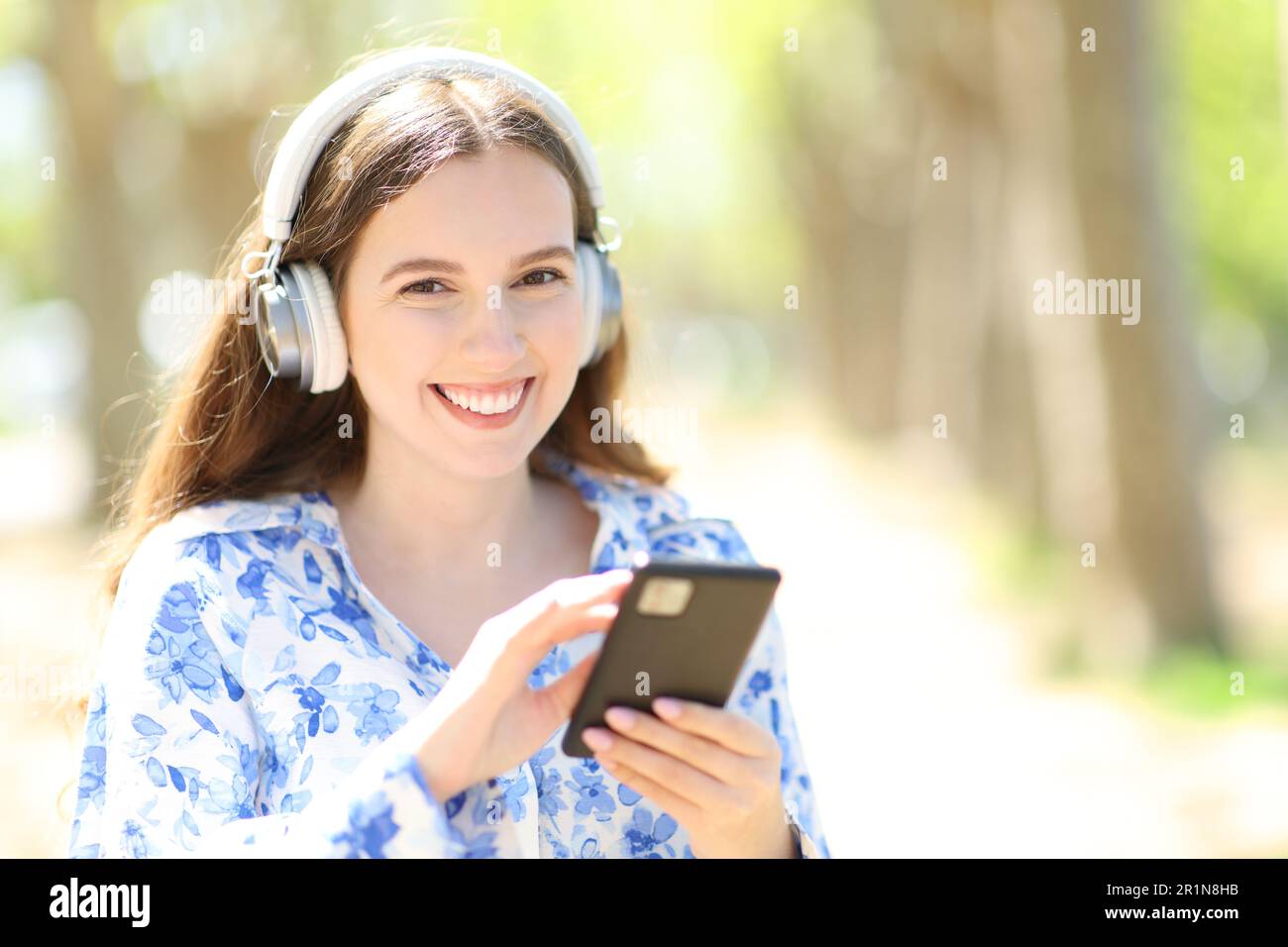 Happy woman wearing headphone listening music holding smartphone looking at camera outdoors Stock Photo