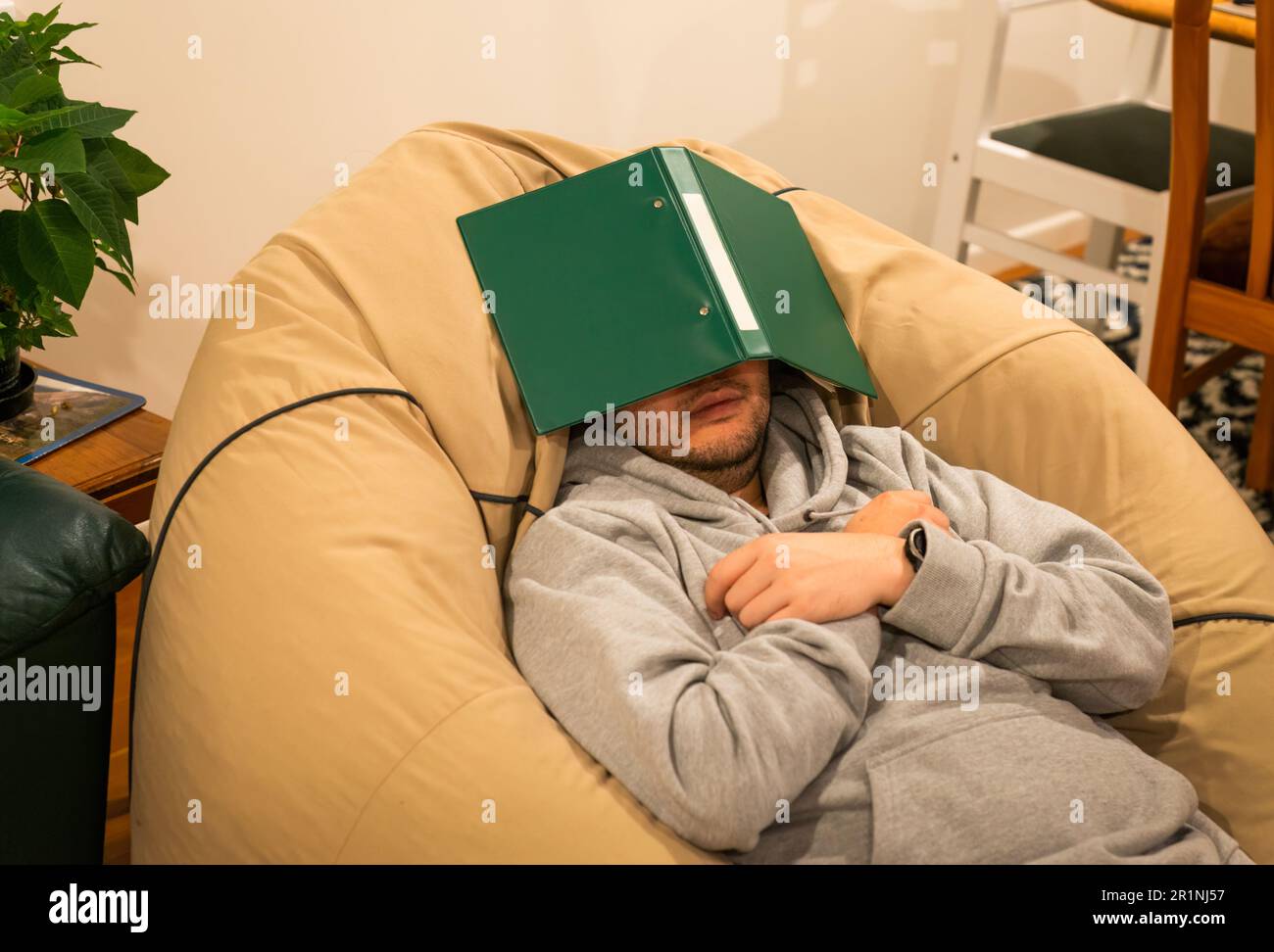Young man lying on bean bag, face covered by green folder. Taking a break from study concepts. Stock Photo