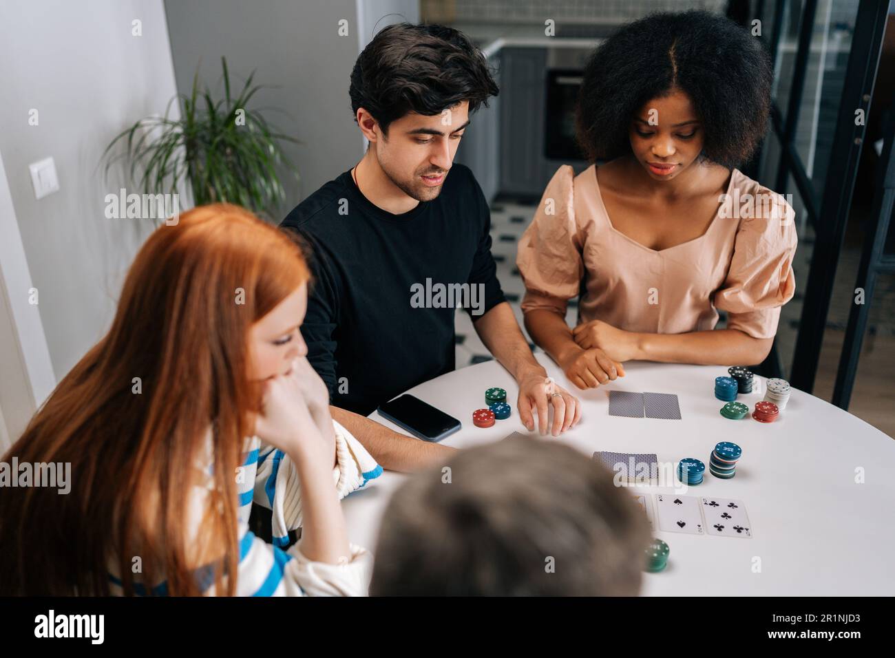 High-angle view of multiethnic friends having home party on weekends playing poker sitting at table in living room. Caucasian and African people Stock Photo
