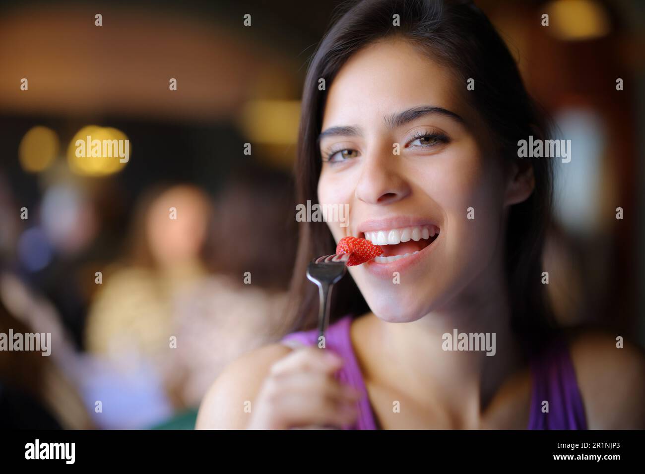 Happy woman eating strawberry in a restaurant interior and looks at camera Stock Photo