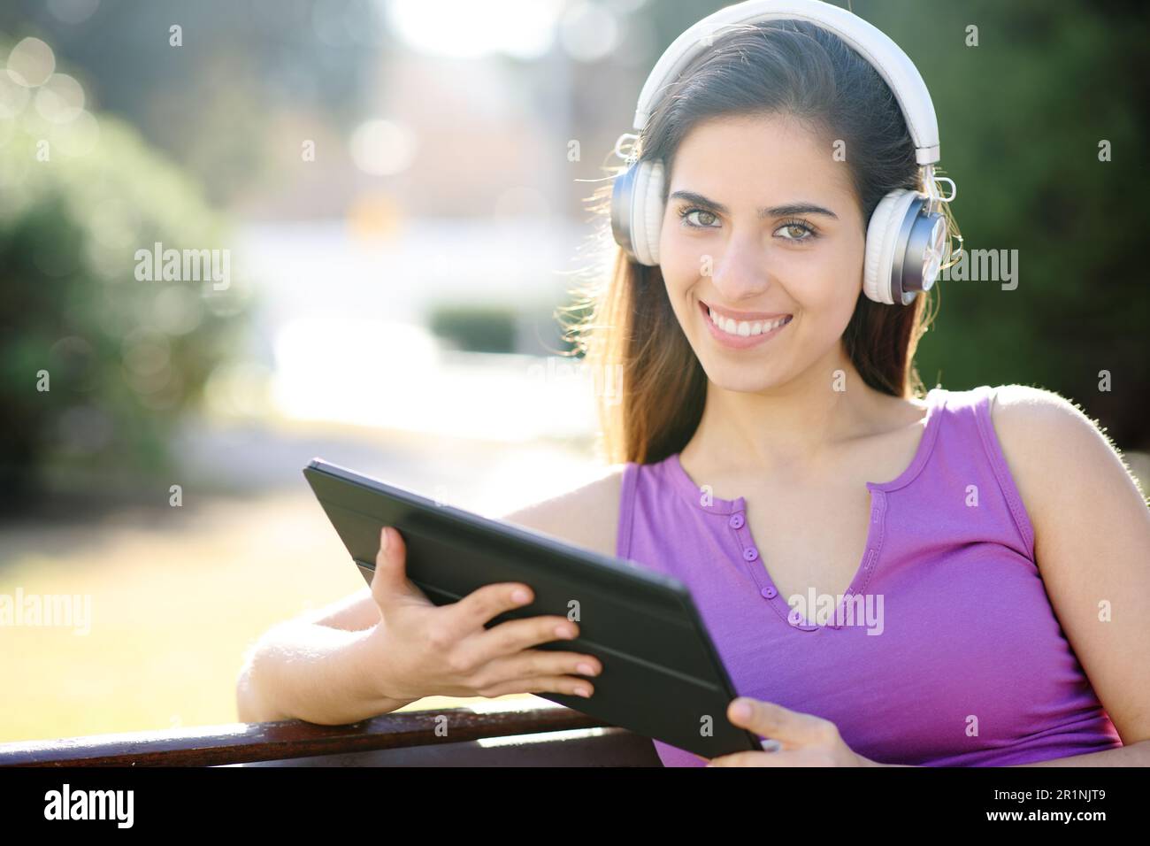 Happy woman with headphone and tablet looks at you sititng in a park Stock Photo