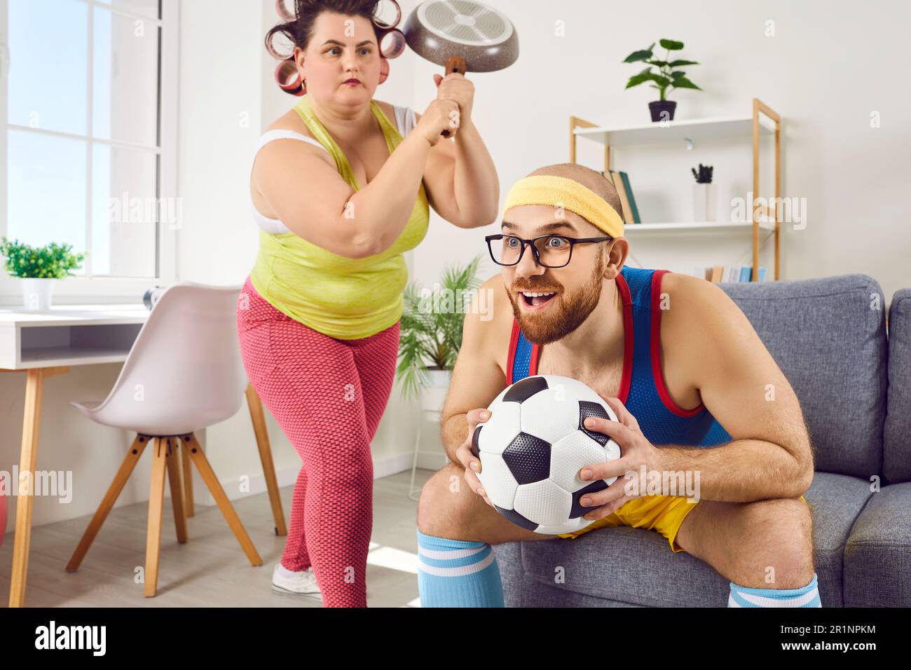 Funny angry housewife holds frying pan over head of her lazy husband watching sport on TV Stock Photo