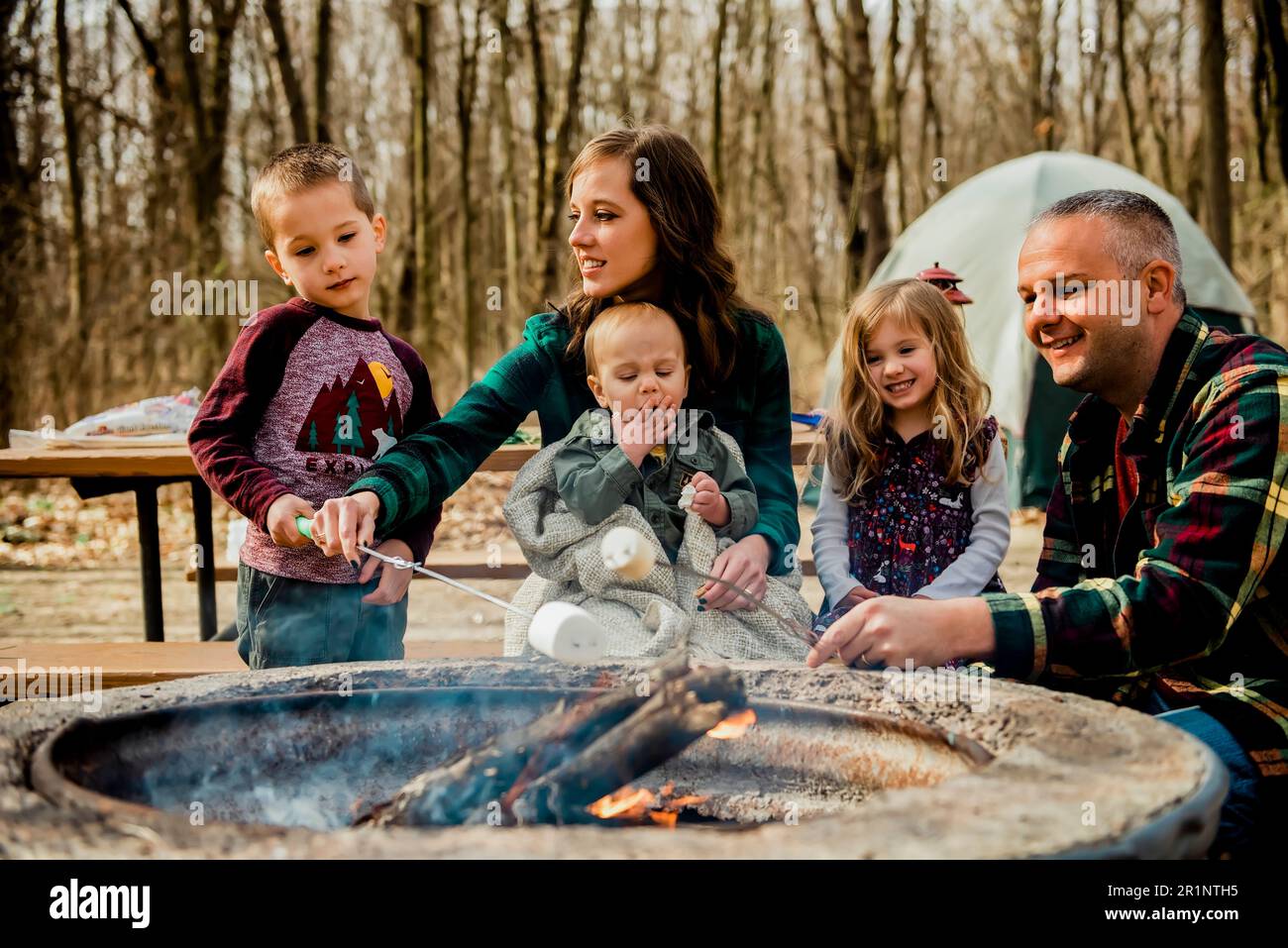 Smiling family roasting marshmallows over campfire in woods during Fal ...