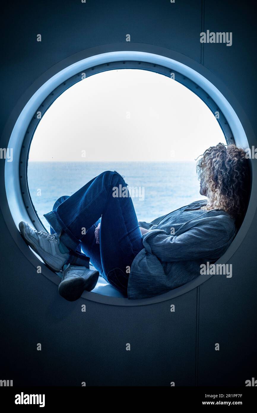 Traveler passenger woman sitting inside a porthole in the boat cruise enjoying the trip and journey travel adventure alone contemplating ocean water. Concept of transport by sea and female people Stock Photo