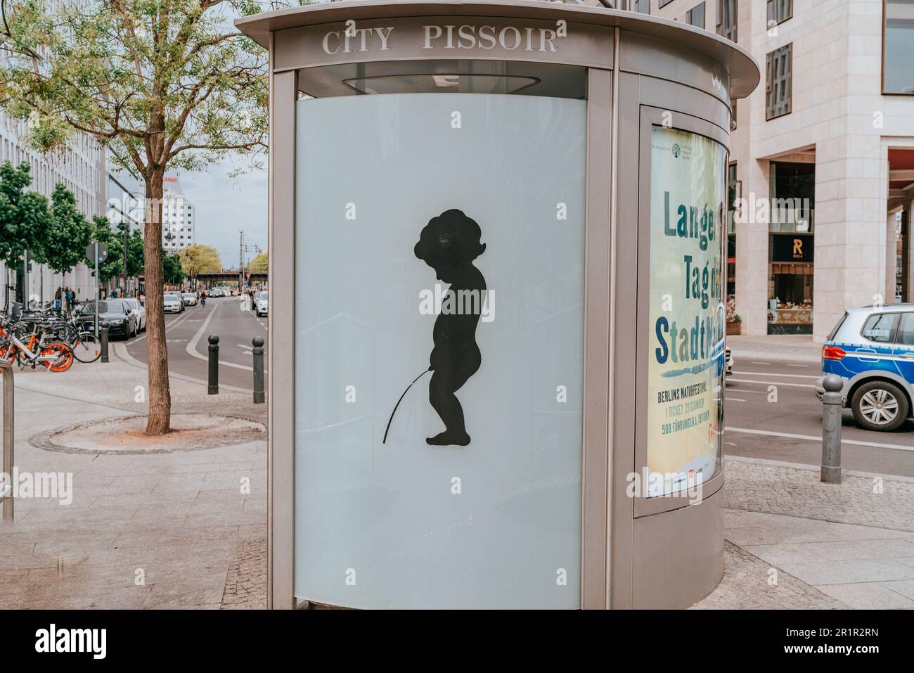 Image captures a public toilet, also known as a city pissoir, situated in a bustling urban