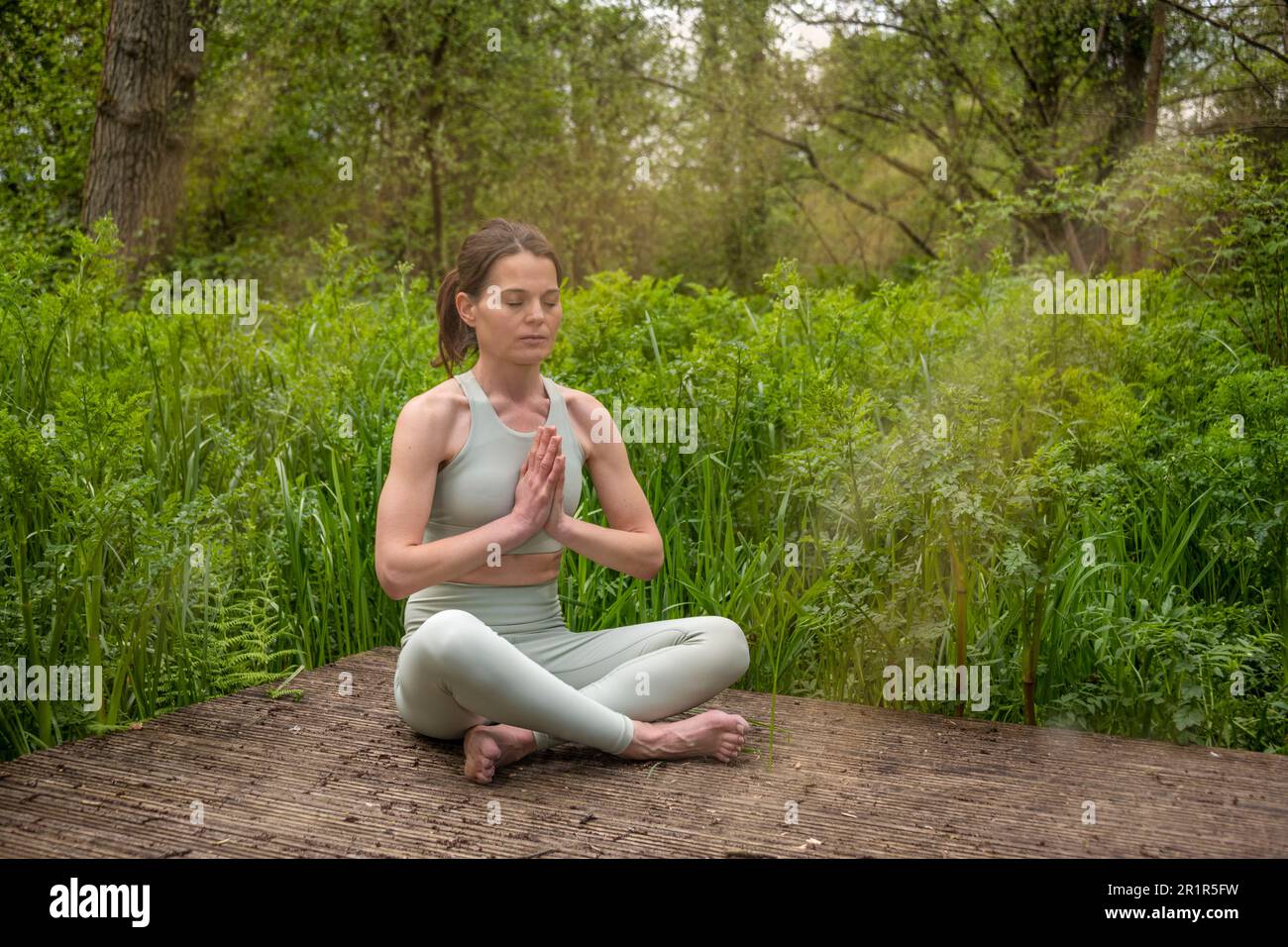 middle aged fit healthy woman practicing yoga outside in natural calm green environment in park meditating with folded palms in front her Stock Photo