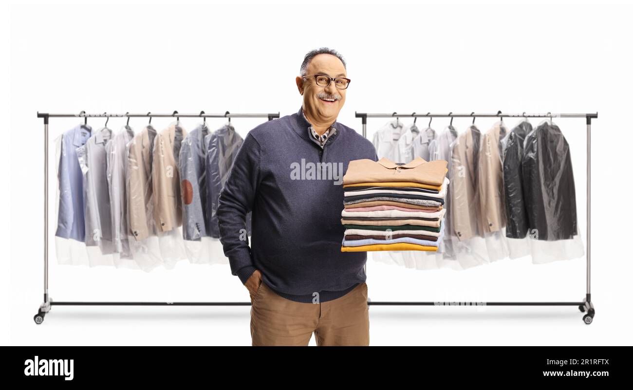 Cheerful mature man holding a pile of folded clothes in front of racks with clothes on hangers isolated on a white background Stock Photo