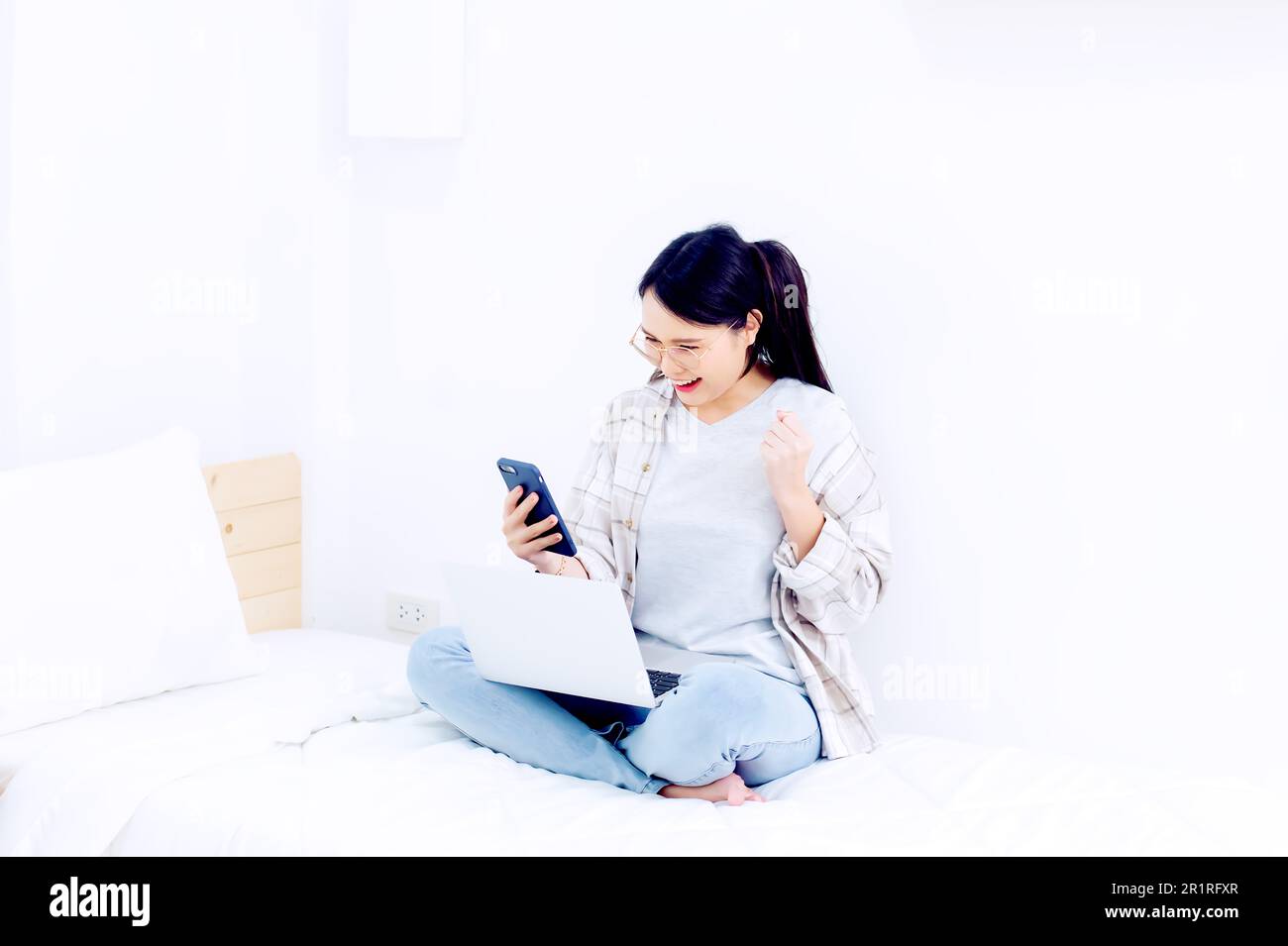 Smiling woman sitting cross-legged on her bed using a mobile phone and laptop computer Stock Photo